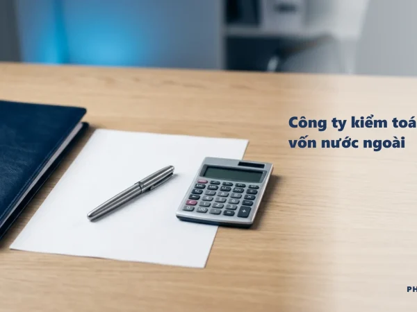 Công ty kiểm toán có vốn nước ngoài: bìa hồ sơ navy, bút ký thép và máy tính để bàn trên giấy kẻ ô